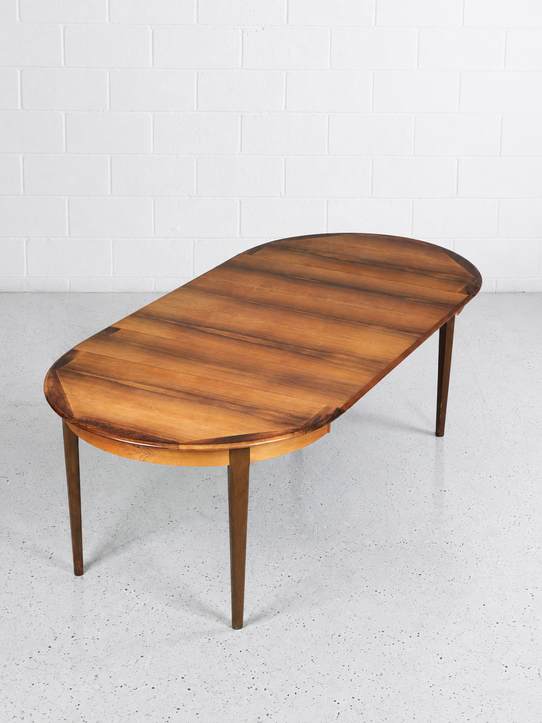 An oval rosewood dining table with a warm medium-brown wood grain finish and tapered legs, set against a white brick wall and light gray concrete floor.