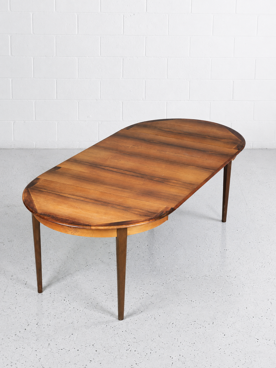 An oval rosewood dining table with a warm medium-brown wood grain finish and tapered legs, set against a white brick wall and light gray concrete floor.